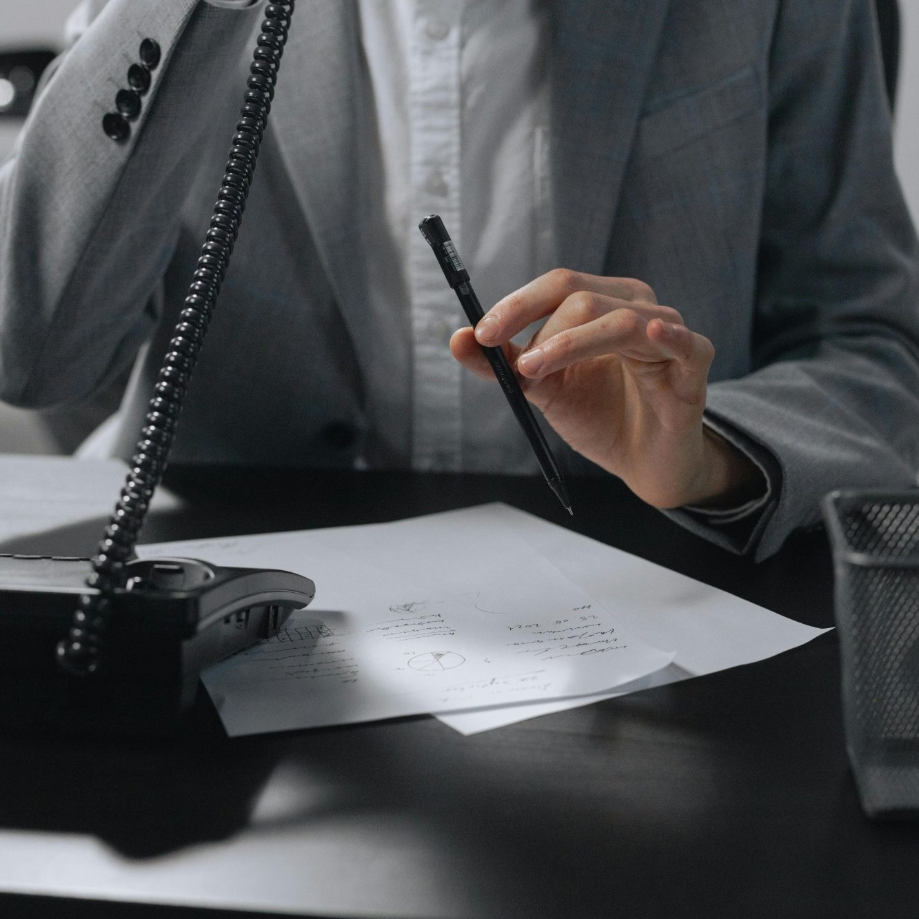 A person in a suit holding a pen near a telephone and documents on a desk.