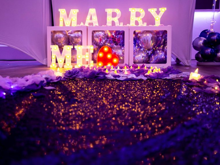 A romantic proposal setup featuring "Marry Me" lights, decorations, and flowers.