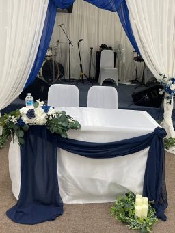 Table decorated with a white cloth and blue drapes, two chairs in front, floral accents.